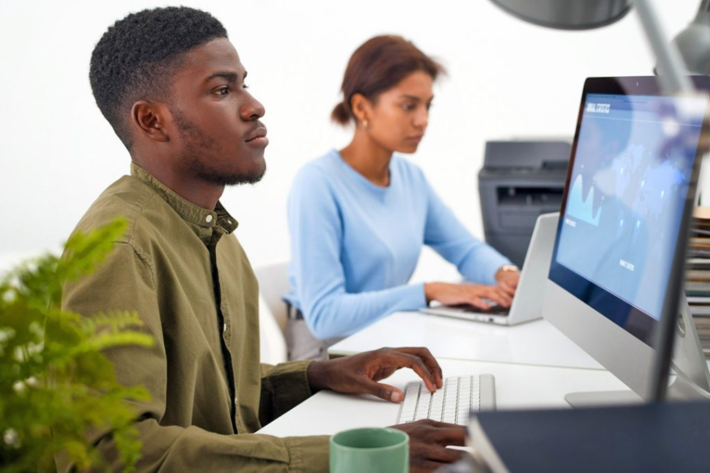 What IT technology should your small business adopt? Young man sitting at his desktop computer in a small office with a woman behind him.